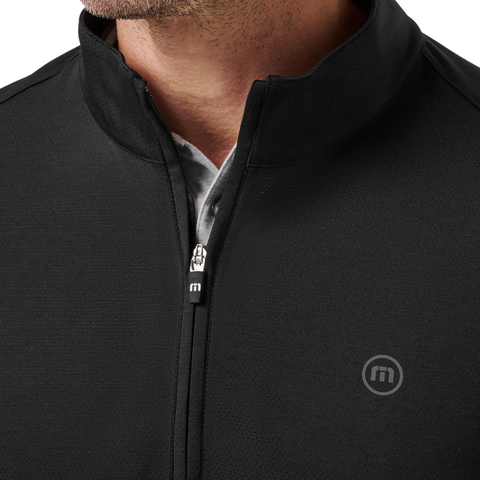 Close-up of a person wearing the Travis Mathew Heater Pro 1/4 Zip Men’s Pullover in black, featuring a lightweight design and a small circular logo. The pullover is partially unzipped to show a light gray collared shirt underneath.