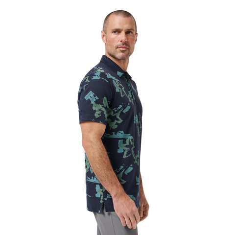 A man with short hair and a trimmed beard wears the Travis Mathew Men’s Favourite Hello Polo in Total Eclipse Blue and light gray pants, standing sideways and looking forward against a plain white background.