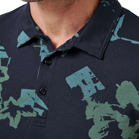 Close-up of a person wearing the TravisMathew Men’s Favourite Hello Polo in Total Eclipse Blue, featuring green and blue abstract patterns, three buttons undone at the collar, and only the lower face and neck visible.
