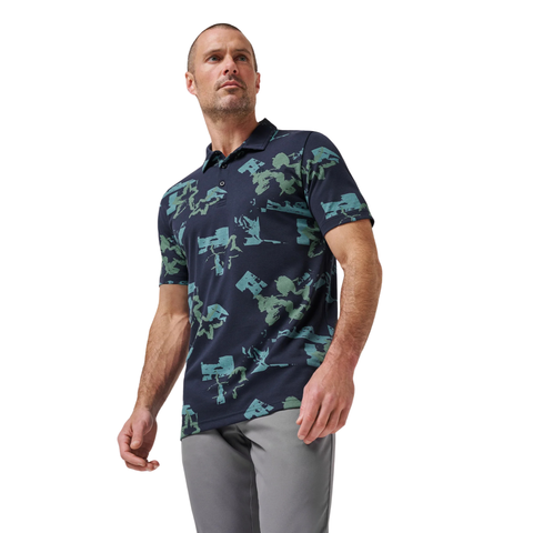 A man wearing a Travis Mathew Men’s Favourite Hello Polo in Total Eclipse Blue and light gray pants stands against a white background, looking slightly to the side.