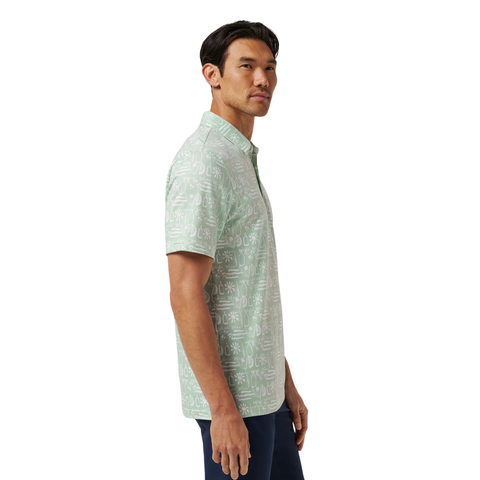 A man stands sideways in the TravisMathew Men’s Featherweight Cabana Polo in Aqua Foam, an ultra-light golf shirt with a white tropical pattern, paired with dark pants against a plain white background.