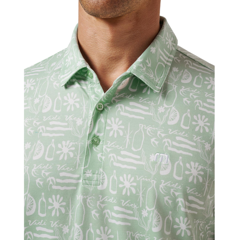 A person wears the TravisMathew Men’s Featherweight Cabana Polo in Aqua Foam, a light green shirt with white floral and abstract patterns and "Vidi Vici" text. The image highlights the collar and upper chest.