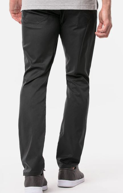 A person stands with their back to the camera, wearing Travis Mathew Men's Open to Close Pants in black, paired with a light gray shirt and gray sneakers, set against a plain white background.