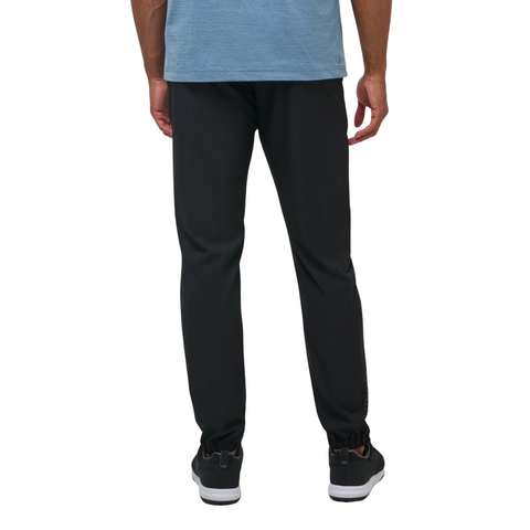 A person is shown from the back wearing a blue short-sleeve shirt, black Travis Mathew Open To Close Men’s Jogger pants, and black sneakers, standing against a plain white background for all-day comfort.