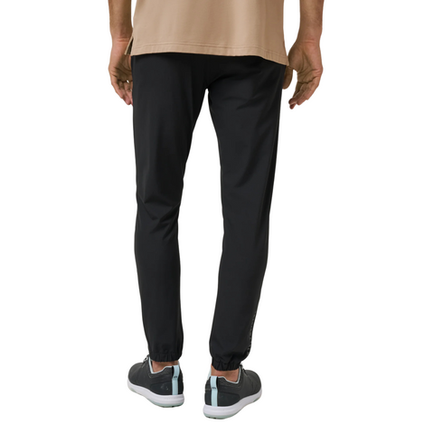 A person in a beige shirt, gray sneakers, and Travis Mathew Open To Close Men’s Jogger in black stands with their back to the camera against a white background, highlighting all-day comfort.
