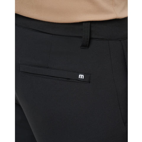 A close-up shows a person wearing the Travis Mathew Open To Close Men’s Jogger in black, featuring a welt pocket and metallic M logo, paired with a beige top for all-day comfort.