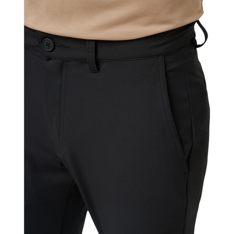 A close-up shows a person wearing Travis Mathew Open To Close Men’s Jogger in black—mens golf joggers with button and zipper closure. A beige shirt is tucked at the waist for comfort. Only the lower torso and upper legs are visible.
