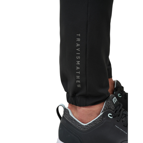 A close-up of a person’s lower leg wearing the TravisMathew Open To Close Men’s Jogger in black, paired with black athletic shoes with white soles and gray laces for all-day comfort.