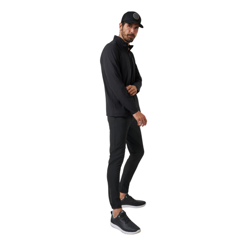 A man wears black athletic wear, featuring the Travis Mathew Open To Close Men’s Jogger, along with a matching jacket, sneakers, and cap, standing slightly turned against a white background for all-day comfort.