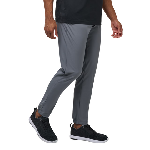 A person wearing Travis Mathew Open To Close Mens Jogger pants in grey stands against a white background, paired with a black short-sleeve shirt and black running shoes. Only the lower body is visible.