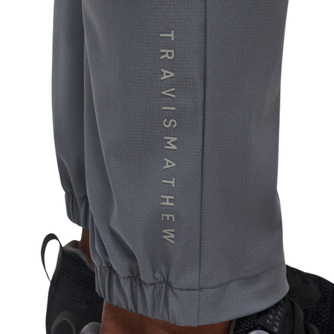 Close-up of the Travis Mathew Open To Close Mens Jogger, featuring TRAVISMATHEW printed vertically on the lower leg. These gray joggers have a tapered fit with elastic cuffs and are shown paired with black sneakers.