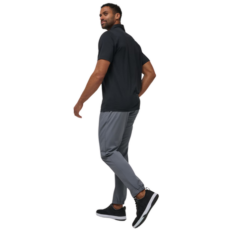 A man in a short-sleeve black polo, TravisMathew Open To Close Mens Jogger pants by Travis Mathew, and black sneakers walks confidently, glancing over his shoulder against a white background.