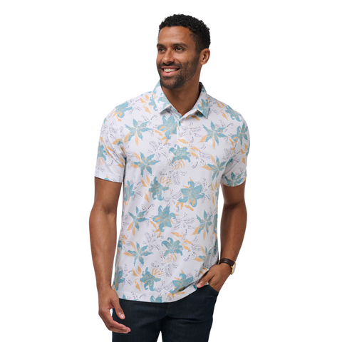 A man wearing the Travis Mathew Paradise Peak Men’s Polo, a short-sleeve golf shirt with a blue, green, and yellow floral design, stands smiling with one hand in his pocket and a watch on his wrist.