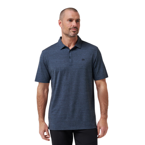 A man with short hair and a beard wears the TravisMathew The Heater Mens Polo by Travis Mathew in dark blue with a subtle pattern and small logo, standing slightly turned and smiling softly in this breathable performance golf polo.