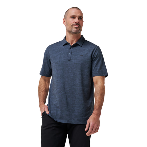 A man with short hair and a trimmed beard wears the Travis Mathew The Heater Mens Polo, made from breathable performance fabric, paired with black pants, standing with one hand in his pocket against a plain white background.