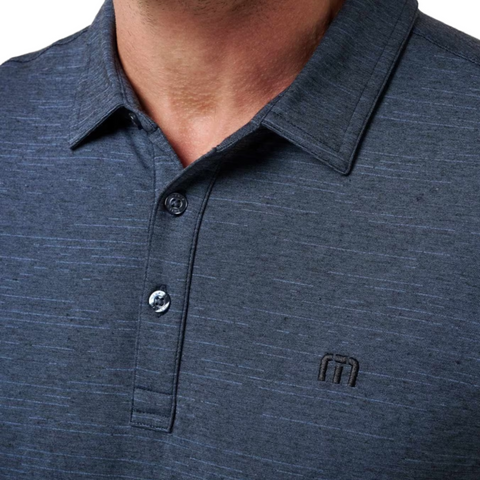 Close-up of a person wearing the TravisMathew The Heater Mens Polo by Travis Mathew in dark blue, with short sleeves, a collar, three buttons, and an embroidered logo on the left chest. Face not visible. This golf polo is breathable for comfort and style.