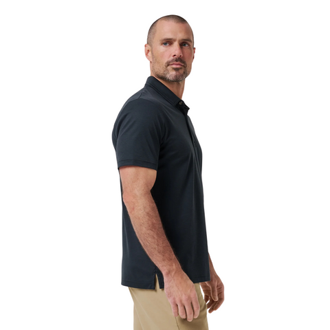A man with short hair and a beard stands sideways, wearing the Travis Mathew The Zinna 2.0 Men’s Polo in black and beige pants, looking slightly toward the camera against a plain white background.