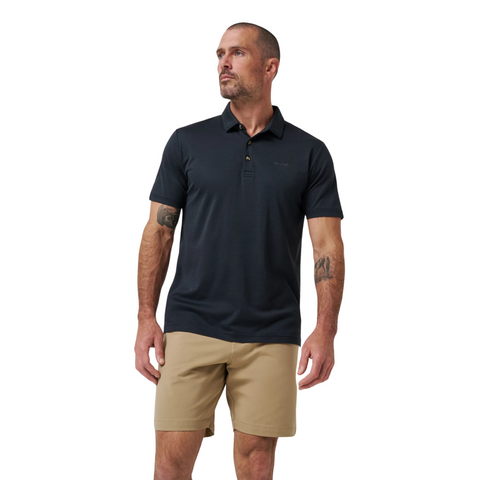 A man with short hair and a beard wears the Travis Mathew The Zinna 2.0 Men’s Polo in black and tan shorts, standing against a white background with his head turned slightly to the side.