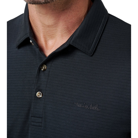 Close-up of a man wearing the Travis Mathew The Zinna 2.0 Men’s Polo in black, featuring subtle horizontal stripes, two-button placket, and small cursive TravisMathew embroidery on the chest.