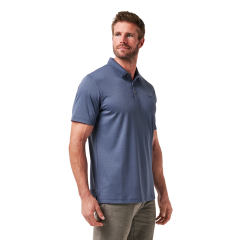 A man with short brown hair and a beard wears the TravisMathew The Zinna 2.0 Men’s Polo in Mood Indigo and gray pants, standing against a white background and looking slightly to the side.