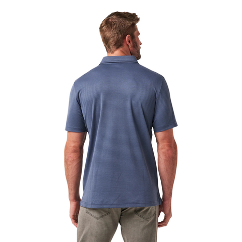 A man faces away from the camera in gray jeans and a Travis Mathew The Zinna 2.0 Men’s Polo in Mood Indigo, set against a plain white background to highlight the stylish design of this performance polo.