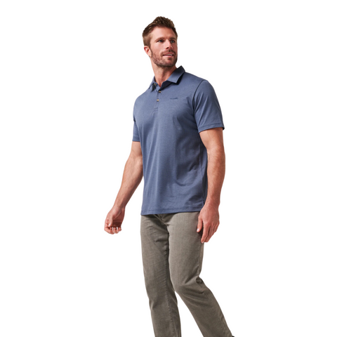 A man with short brown hair and a beard stands against a plain white background, wearing light gray pants and the Travis Mathew The Zinna 2.0 Men’s Polo in Mood Indigo, looking slightly to his left.