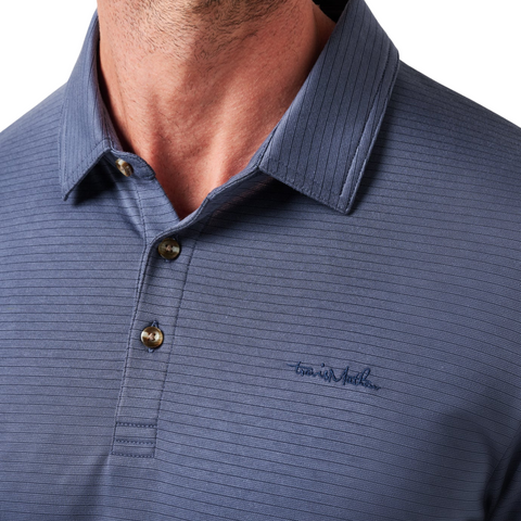 A close-up of a man wearing the Travis Mathew The Zinna 2.0 Men’s Polo in Mood Indigo, a blue short-sleeve button-up with subtle stripes and embroidered text on the chest, cropped from chin to chest.