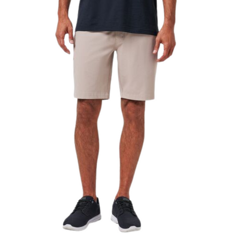 A person, visible from the waist down, wears Travis Mathew Wanderlust Mens Shorts in light beige, paired with a dark navy T-shirt and black athletic shoes, standing against a plain white background.