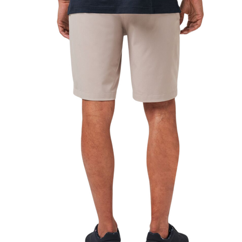 Seen from the back, a person wears Travis Mathew Wanderlust Mens Shorts in beige with a dark shirt and black sneakers. The lightweight shorts stand out against the plain white background.