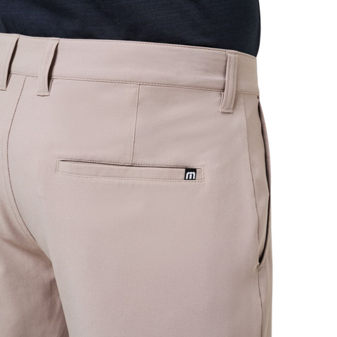 A close-up of the back of a person wearing Travis Mathew Wanderlust Mens Shorts in beige, featuring a small black and white m logo tag above the back pocket. A dark shirt is tucked into these lightweight golf shorts.