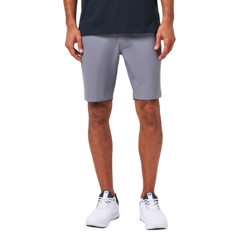A person, visible only from the waist down, wears a dark shirt, white sneakers, and light gray Travis Mathew Wanderlust Mens Shorts while standing against a plain white background.