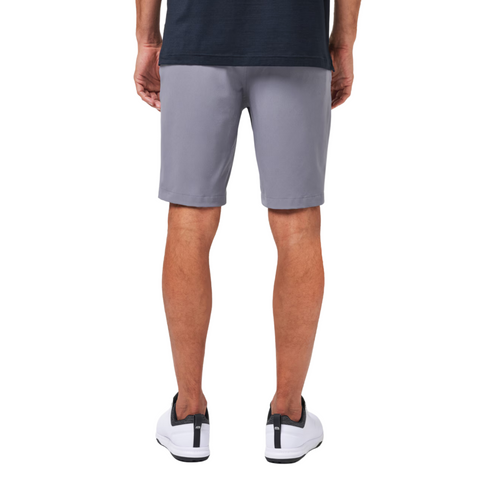 A person stands with their back to the camera, wearing a navy shirt, Travis Mathew Wanderlust Mens Shorts in light gray, and white sneakers with black accents.