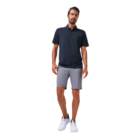 A man stands against a white background wearing a dark navy polo, Travis Mathew Wanderlust Mens Shorts in light gray, and white athletic shoes, showcasing stylish and lightweight shorts ideal for any casual look.