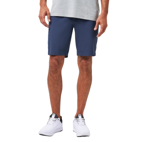 A person wearing a gray t-shirt, Travis Mathew Wanderlust Mens Shorts in navy blue, and white athletic shoes stands against a plain white background. The photo is cropped from the shoulders down.