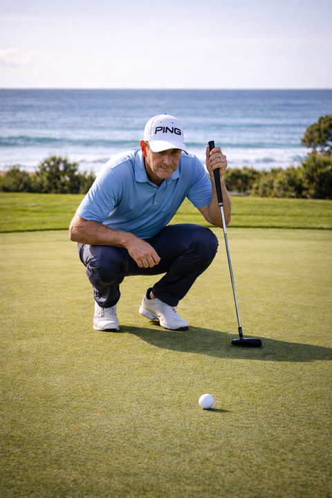 A golfer wearing a ping cap and with the ocean in the background is crouched down to read his putt on the putting green