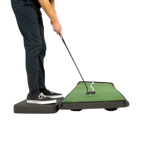 A person in black pants and sneakers practices putting on an elevated indoor putting green mat with a golf club and a golf ball aimed at a hole.