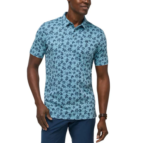 A man wearing a light blue short-sleeve button-up shirt with a dark blue palm tree pattern, paired with navy pants and a brown watch, stands smiling with one hand in his pocket.