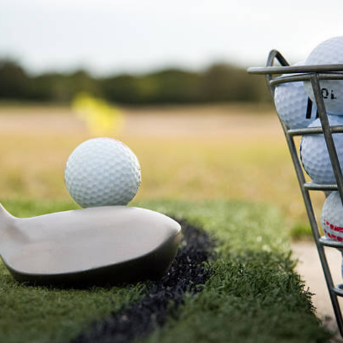 A close-up of a golf club about to hit a golf ball on artificial turf, with a basket of golf balls nearby and a blurred outdoor background.