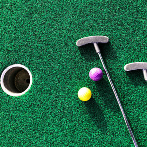 Two mini golf putters and a yellow and a purple golf ball sit on artificial green turf near a hole.