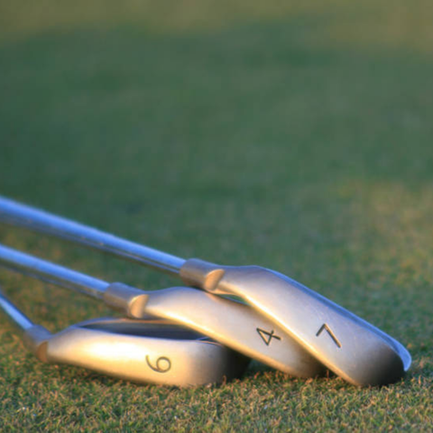 Three golf irons numbered 6, 4, and 7 lay on green grass, overlapping each other with their clubheads resting on the ground and shafts extending out of the frame.