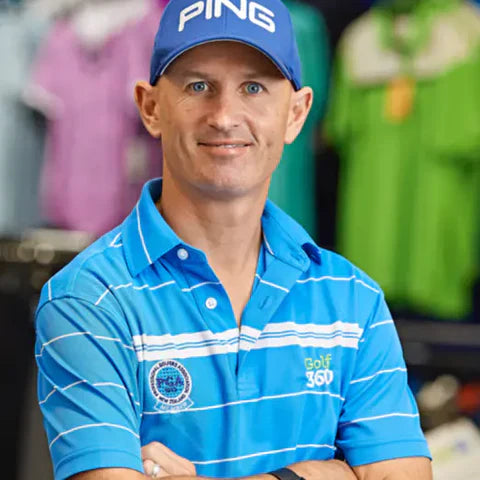 A man wearing a blue PING cap and a blue striped Golf 360 polo shirt stands with his arms crossed, smiling in a store with colorful shirts in the background.