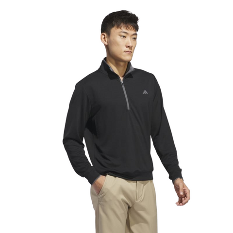 A man wearing the Adidas Mens Core Lightweight Zip Top in black and beige pants stands with one hand in his pocket against a white background.