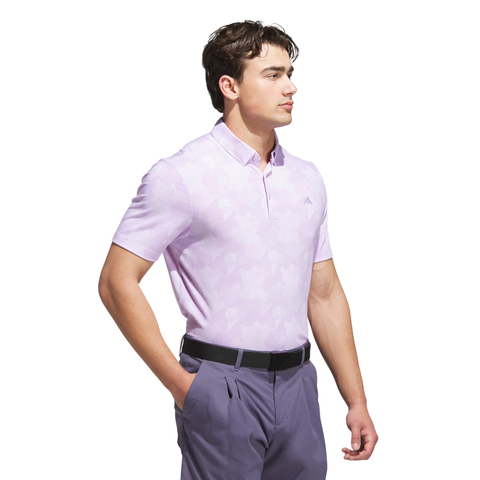 A man stands against a white background, looking to the side with one hand in his pocket, wearing an Adidas Men's Go-To Printed Polo Shirt in Powder Plum/White tucked into gray pants with a black belt.