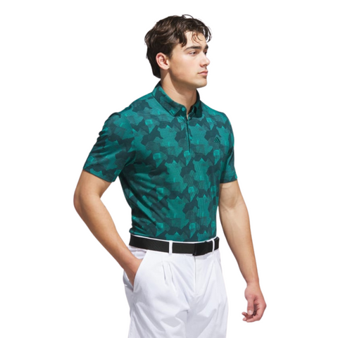A man wearing an Adidas Mens Go-To Printed Polo Shirt in Pure Teal/Ivy stands with his hands in his pockets, paired with white pants and a black belt, looking to the side against a white background.