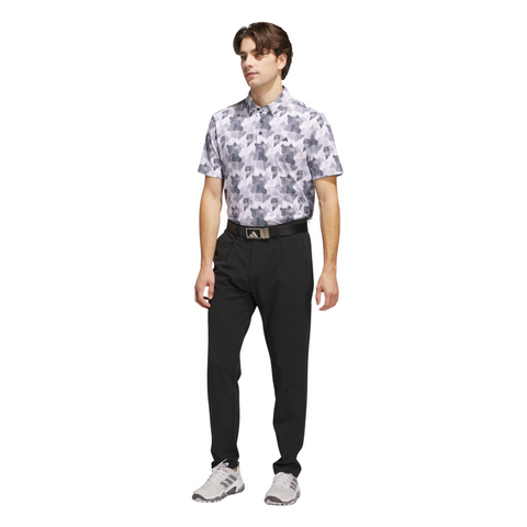 A man wearing the Adidas Men's Go-To Printed Polo Shirt in White/Collegiate Navy, paired with black pants, a black belt, and gray sneakers, stands against a plain white background.