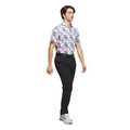 A man with brown hair wears the Adidas Men's Go-To Printed Polo Shirt in White/Collegiate Navy, paired with black pants, a black belt, and gray sneakers as he walks against a white background.