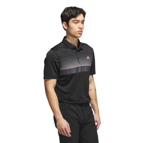 A young man wears the Adidas Men's Performance Allover Print Polo Shirt in black, featuring a gray and pink patterned stripe across the chest, paired with black pants, standing against a white background.