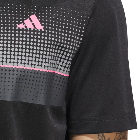 Close-up of a person wearing the Adidas Men's Performance Allover Print Polo Shirt in black, featuring a pink logo and a pattern of white, gray, and pink dots and lines on the chest. Part of the tattooed arm is visible.