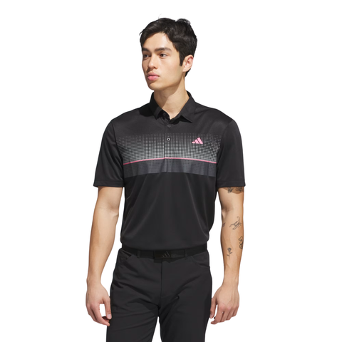 A man stands against a white background wearing the Adidas Men's Performance Allover Print Polo Shirt in black, featuring a dotted pattern and pink chest accents, paired with black pants and a belt.