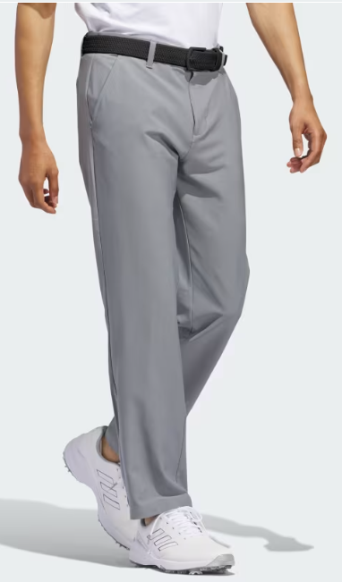 A person wearing Adidas Mens Ultimate 365 Trousers Grey, paired with a black belt and white athletic shoes, stands with one leg crossed in front of the other against a plain background.